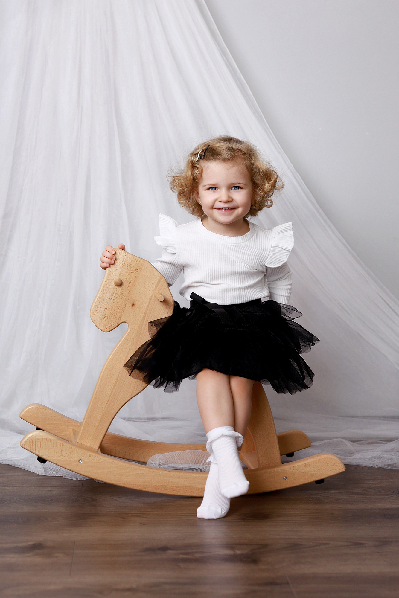 kislány elegáns ruhában hintalovon pózol mosolyogva.