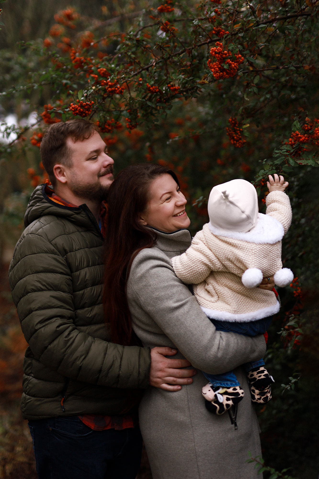 Apuka és anyuka áll a kisbabával a kezükben egy kültéri fotózáson egy narancssárgabogyós bokor előtt.